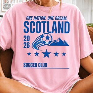 May include: A light pink t-shirt featuring blue text and graphics. The design includes the words ONE NATION. ONE DREAM. SCOTLAND 2026 SOCCER CLUB, a soccer ball in motion, a mountain silhouette, and a row of five blue stars centered on the chest.
