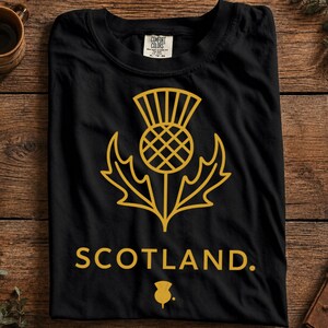 May include: A black short-sleeved t-shirt featuring a gold-colored line art graphic of a Scottish thistle flower centered on the chest. Below the thistle, the word SCOTLAND is printed in gold capital letters, followed by a small thistle icon.