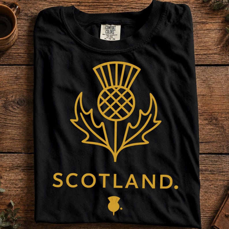 May include: A black short-sleeved t-shirt featuring a gold-colored line art graphic of a Scottish thistle flower centered on the chest. Below the thistle, the word SCOTLAND is printed in gold capital letters, followed by a small thistle icon.