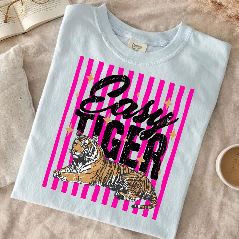 Pu&ograve; includere: T-shirt azzurra con una grafica di tigre e la scritta "Easy Tiger". Il design include strisce verticali rosa e stelle dorate. La maglietta &egrave; realizzata in un materiale morbido.
