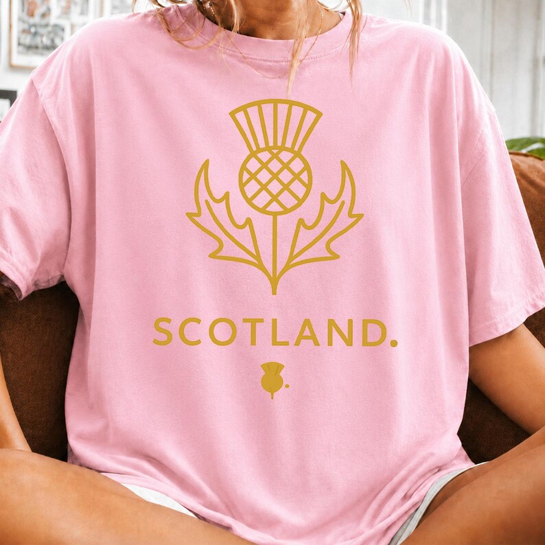May include: A light pink short-sleeve t-shirt featuring a gold-colored graphic of a thistle flower centered on the chest. Below the thistle, the word SCOTLAND is printed in gold capital letters, followed by a small gold thistle icon and a period.