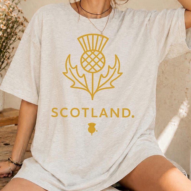 May include: A light heather gray short-sleeve t-shirt featuring a gold-colored graphic of a stylized Scottish thistle flower. Below the thistle, the word SCOTLAND is printed in gold capital letters, followed by a small gold thistle icon.