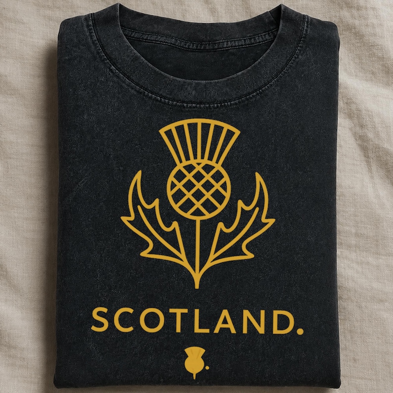 May include: A black crew neck t-shirt featuring a gold graphic of a thistle flower centered on the chest. Below the thistle, the word SCOTLAND is printed in gold capital letters, followed by a small, simplified gold thistle icon at the bottom.