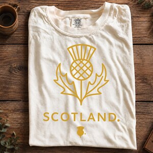 May include: An off-white short-sleeve t-shirt featuring a gold-colored graphic of a Scottish thistle flower centered on the chest. Below the thistle, the word SCOTLAND is printed in gold capital letters, followed by a small thistle icon.