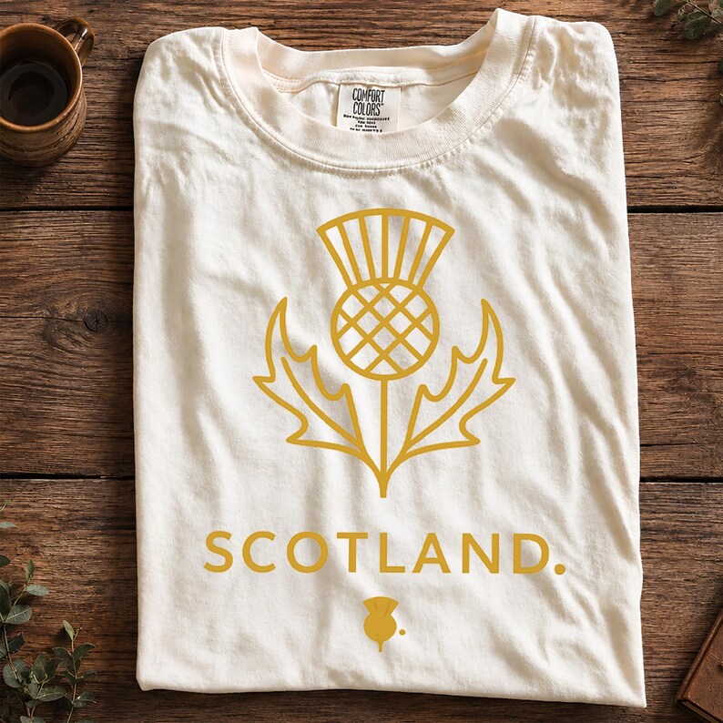 May include: An off-white short-sleeve t-shirt featuring a gold-colored graphic of a Scottish thistle flower centered on the chest. Below the thistle, the word SCOTLAND is printed in gold capital letters, followed by a small thistle icon.