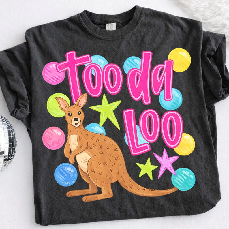 K&ouml;nnte beinhalten: Schwarzes T-Shirt mit einem Cartoon-K&auml;nguru und dem pinkfarbenen Text "Tooda Loo". Das Design umfasst bunte Sterne und Blasenakzente in Pink, Gelb, Gr&uuml;n und Blau. Das Shirt ist aus weichem Material.