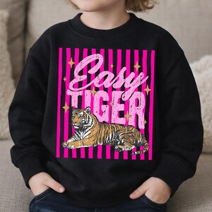 Pu&ograve; includere: Felpa nera con un design a righe rosa e nere e la scritta "Easy Tiger" in un font retr&ograve;. Il design include un'illustrazione dettagliata di una tigre in marrone e nero, centrata sotto il testo.