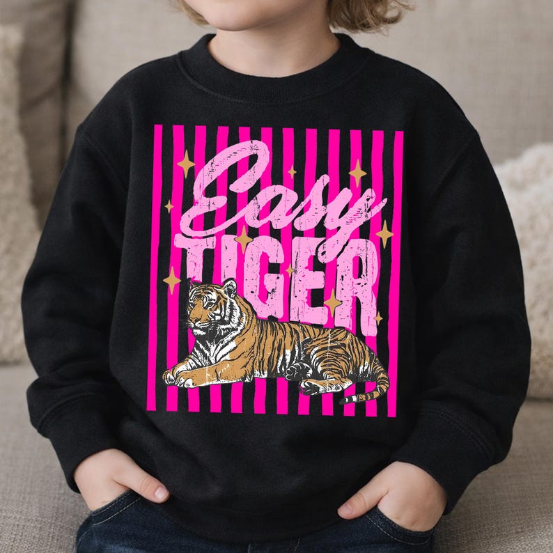 Pu&ograve; includere: Felpa nera con un design a righe rosa e nere e la scritta "Easy Tiger" in un font retr&ograve;. Il design include un'illustrazione dettagliata di una tigre in marrone e nero, centrata sotto il testo.