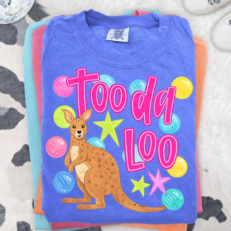 K&ouml;nnte beinhalten: Ein blaues T-Shirt mit den pinkfarbenen Worten "Tooda Loo", das ein Cartoon-K&auml;nguru, Sterne und bunte Blasen zeigt. Das Shirt ist Teil eines Stapels anderer bunter Shirts.