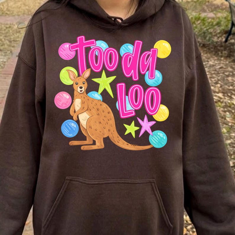 K&ouml;nnte beinhalten: Ein brauner Kapuzenpullover mit einer K&auml;nguru-Illustration und dem Text "Tooda Loo" in Pink und Wei&szlig;. Das Design umfasst bunte Sterne und Tupfen in Pink, Gelb und Blau. Der Hoodie hat eine Vordertasche und eine Kapuze mit Kordelzug.