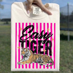 Pu&ograve; includere: Felpa color crema con un design grafico. Il design presenta le parole "Easy Tiger" in nero, su uno sfondo a righe rosa. Un'illustrazione di tigre &egrave; sotto il testo, con accenti di stelle dorate.