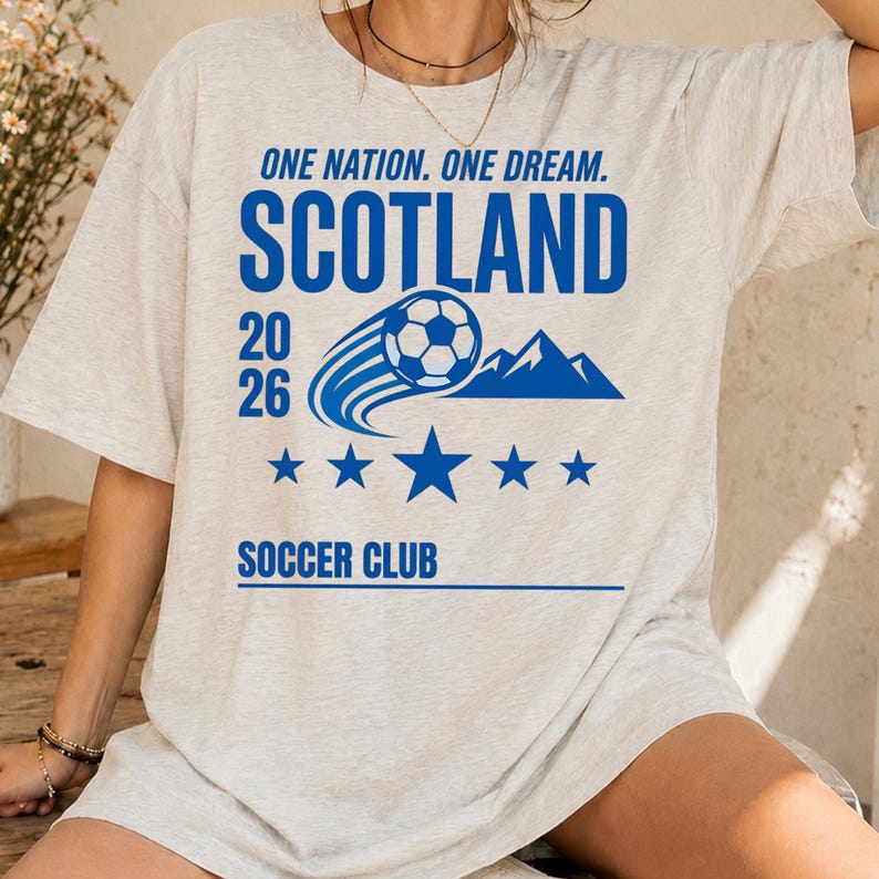 May include: A light heather gray t-shirt featuring blue text that reads ONE NATION. ONE DREAM. SCOTLAND 20 26 SOCCER CLUB. The design includes a graphic of a soccer ball in motion, a mountain silhouette, and a row of five blue stars centered below.