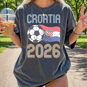 May include: A charcoal gray short-sleeve t-shirt featuring a graphic print with the text CROATIA at the top, a soccer ball, a stylized Croatian flag, and the year 2026 at the bottom in a retro, multi-colored layered font design.