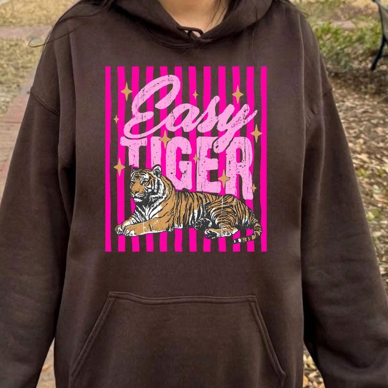 Pu&ograve; includere: Una felpa con cappuccio marrone con una grafica rosa e bianca che dice "Easy Tiger". La grafica presenta un'illustrazione di una tigre. La felpa con cappuccio ha una tasca frontale e un cappuccio con coulisse.
