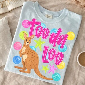 K&ouml;nnte beinhalten: Hellblaues T-Shirt mit einem Cartoon-K&auml;nguru und den Worten "Too da Loo" in Pink. Das Design umfasst bunte Sterne und blasenartige Formen in Pink, Blau und Gelb. Das Shirt ist aus einem weichen Material.