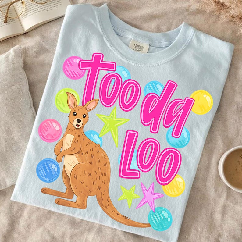 K&ouml;nnte beinhalten: Hellblaues T-Shirt mit einem Cartoon-K&auml;nguru und den Worten "Too da Loo" in Pink. Das Design umfasst bunte Sterne und blasenartige Formen in Pink, Blau und Gelb. Das Shirt ist aus einem weichen Material.