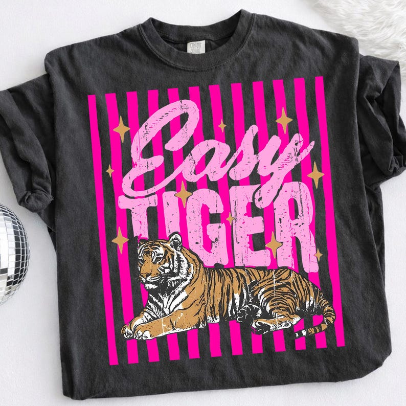 Pu&ograve; includere: T-shirt grigio scuro con un design a righe rosa e bianche. La maglietta presenta la scritta "Easy Tiger" in rosa sopra un'illustrazione dettagliata di una tigre. Accenti di stelle dorate sono sparsi intorno al testo.