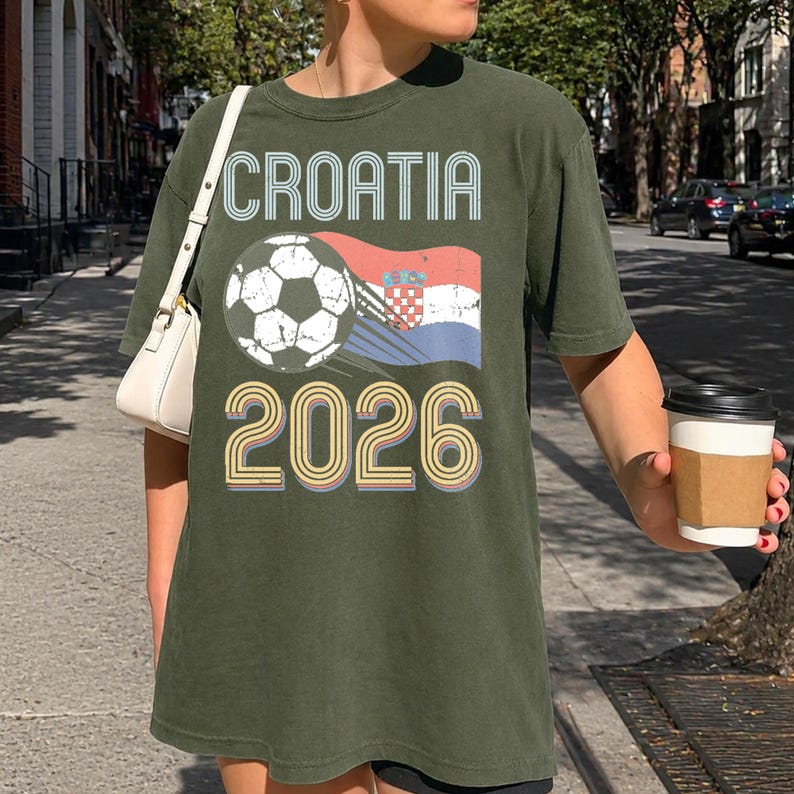 May include: An olive green short-sleeve t-shirt featuring a graphic print with the text CROATIA in a retro font, a soccer ball, a waving Croatian flag, and the year 2026 in large, multi-colored outlined numbers centered on the front chest.