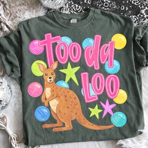 K&ouml;nnte beinhalten: Salbeigr&uuml;nes T-Shirt mit einem Cartoon-K&auml;nguru und dem pinkfarbenen Text "Tooda Loo". Das Design umfasst bunte Blasen- und Sternakzente. Das T-Shirt ist aus weichem Material.