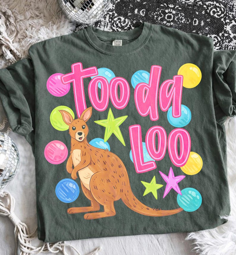 K&ouml;nnte beinhalten: Salbeigr&uuml;nes T-Shirt mit einem Cartoon-K&auml;nguru und dem pinkfarbenen Text "Tooda Loo". Das Design umfasst bunte Blasen- und Sternakzente. Das T-Shirt ist aus weichem Material.
