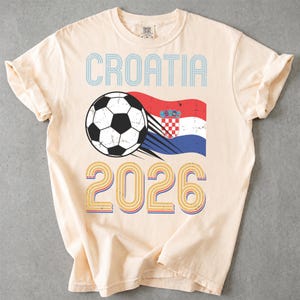 May include: A cream-colored short-sleeve t-shirt featuring the text CROATIA in blue, a black and white soccer ball graphic with motion lines, the Croatian flag, and the year 2026 in a multi-colored layered font design on the front center.