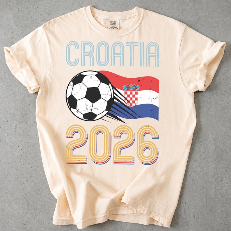 May include: A cream-colored short-sleeve t-shirt featuring the text CROATIA in blue, a black and white soccer ball graphic with motion lines, the Croatian flag, and the year 2026 in a multi-colored layered font design on the front center.