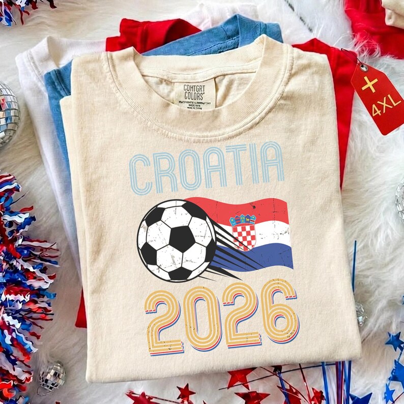 May include: A beige short-sleeved t-shirt featuring the text CROATIA in light blue, a black and white soccer ball, a waving Croatian flag, and the year 2026 in yellow and blue outline. The shirt has a Comfort Colors brand label at the neck.