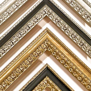May include: A collection of three nested picture frame corners arranged in a chevron pattern. The top frame is silver with ornate floral carvings, the middle is gold with similar detailing, and the bottom is a black frame with gold accents.