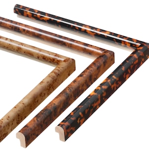 May include: Close-up of various wooden picture frame samples. The frames feature different wood grains and finishes, including a tortoiseshell pattern and a light-colored burl wood. The frames are cut at a 45-degree angle for corner assembly.