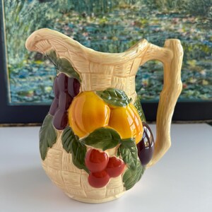 May include: A ceramic pitcher with a basket weave design. The pitcher is decorated with a cluster of oranges, cherries, and plums. The handle is shaped like a tree branch. The colors are beige, orange, red, green, and purple.