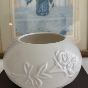 May include: A white ceramic vase with a round shape and a wide opening. The vase features embossed floral designs, including roses and leaves. The vase is sitting on a dark brown surface.