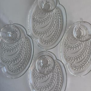 May include: Four clear glass teacup and saucer sets. Each set features an oval-shaped saucer with a raised, leaf-like pattern and a matching teacup. The teacups sit in a circular indentation on the saucers.