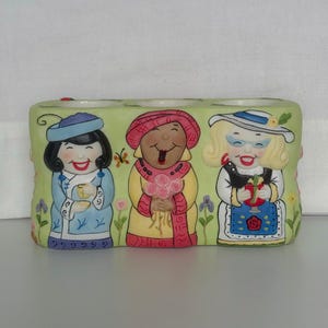 May include: A rectangular ceramic planter featuring three smiling figures in traditional attire. The figures are set against a light green background with floral accents. The planter is painted with bright colors and has a whimsical design.