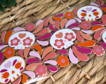 May include: A clear acrylic sheet featuring a repeating pattern of white sugar skulls decorated with pink and orange heart and flower motifs, interspersed with various pink, orange, and magenta petal-shaped designs across the surface.