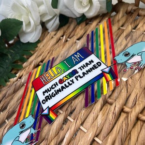 May include: A clear acrylic name tag pin featuring a rainbow-colored background with diagonal stripes. The text reads, "HELLO, I AM MUCH GAYER THAN ORIGINALLY PLANNED." Two light blue frogs are positioned on the sides, each holding a small pride flag.