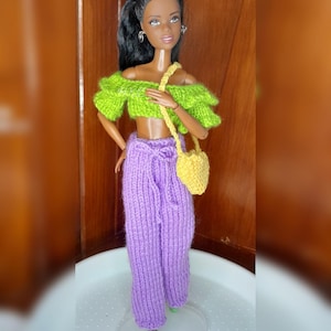 May include: A doll wearing a lime green off-the-shoulder top, purple knit pants, and a yellow purse. The doll has black hair and is standing on a clear stand. The outfit is handmade.