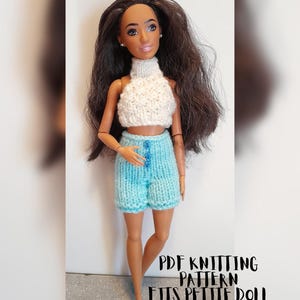 May include: A doll wearing a handmade knitted outfit. The outfit includes a white turtleneck crop top and light blue shorts with blue buttons. The doll has long brown hair and is wearing blue shoes. Text on the image reads "PDF Knitting Pattern Fits Petite Doll."