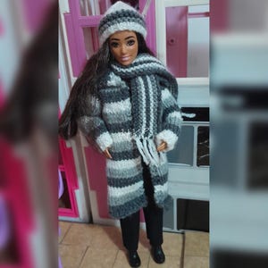 May include: A doll with long dark hair wearing a hand-knitted ensemble consisting of a grey and white striped long coat, a matching striped scarf with fringe, and a grey and white beanie. The doll is also wearing plain black trousers and black shoes.