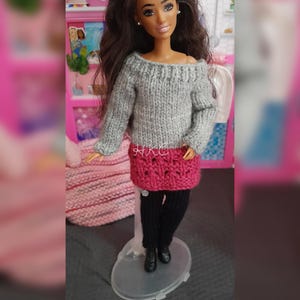 May include: A doll with long dark brown hair wearing a gray off-the-shoulder knit sweater, a dark pink textured knit skirt, and black knit leggings. The doll is standing on a clear plastic display stand and features small white stud earrings.