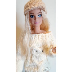 May include: A doll wearing a cream-colored knitted outfit, including a hat, sweater with an owl design, and pants. The doll has blonde hair and blue eyes. The sweater features an off-the-shoulder design. The initials "HKE" are visible on the pants.