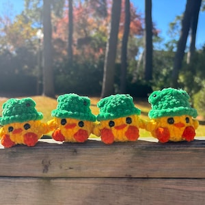 May include: Four handmade crochet ducklings with yellow bodies, orange beaks and feet, and green hats. The ducklings are arranged in a row on a wooden surface, with a blurred background of trees and a blue sky.