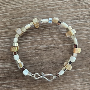 May include: A bracelet featuring a repeating pattern of smooth white beads, irregular tan shell-like chips, and small metallic silver-toned cube spacers. The piece is secured with a silver-toned metal hook and eye clasp closure at the bottom.