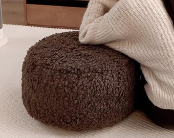 May include: A round, brown, textured ottoman. The ottoman has a plush, shaggy appearance and sits on a white surface. The image is taken from a slightly elevated angle, showcasing the ottoman's shape and texture.