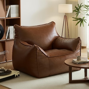 May include: A large, brown leather beanbag chair with a rounded shape and a low profile. The chair has a soft, inviting appearance. A small, round wooden side table and a floor lamp are nearby. The chair is approximately 100 cm wide.