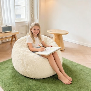 May include: A large, cream-coloured, fluffy beanbag chair on a green rug. A young person is seated in the chair, holding a drawing pad and pencil. A small wooden side table and speaker are visible in the background.