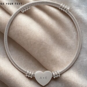 May include: A silver bangle bracelet featuring a heart-shaped charm with the initials "A+E". The bracelet has a twisted cable design. The text "CHOOSE YOUR TEXT" is at the top of the image.