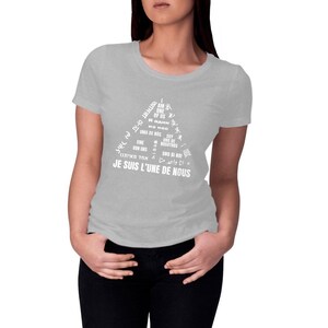Può includere: Una maglietta grigio chiaro con un design triangolare bianco. Il design include la frase "I AM ONE OF US" in più lingue. Il testo "JE SUIS L'UNE DE NOUS" è stampato nella parte inferiore del triangolo.