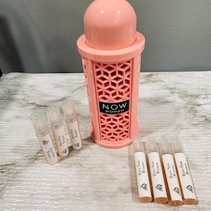 May include: A light pink perfume bottle with a geometric design and the text "NOW WOMEN". Several small perfume vials are next to the bottle. The vials are labeled "Rave Now Woman".