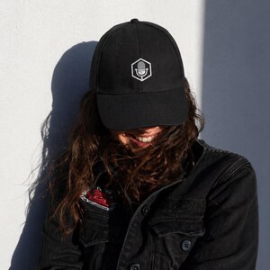 May include: Black baseball cap with a white microphone logo in a hexagon shape. The cap is worn by someone wearing a black denim jacket. The background is a white wall with a shadow.