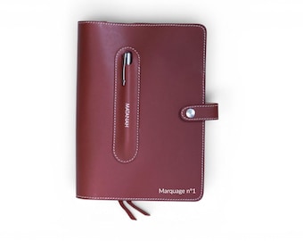 Puede incluir: Una funda de cuaderno de cuero rojo oscuro con costuras blancas, que presenta una ranura vertical para bolígrafo con un bolígrafo plateado, un cierre de botón a presión y el texto "MATANAH" y "Marquage n°1" impreso en blanco en la portada.
